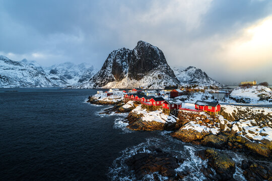 Hamn&oslash;y auf den Lofoten in Norwegen zur goldenen Stunde mit warm beleuchteten Rorbuer-H&auml;usern, w&auml;hrend der Festh&aelig;ltinden im Hintergrund von Wolken umgeben ist und im Schatten liegt