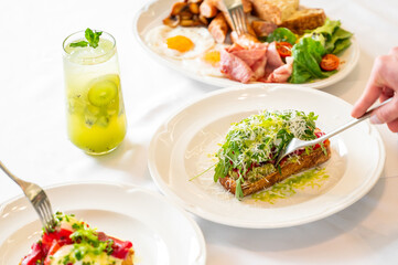 A hand holds a fork ready to enjoy a fresh open-faced sandwich topped with greens and tomatoes. In the background, a refreshing drink and additional breakfast plates complete the meal.