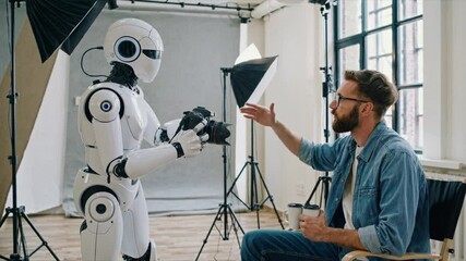 White humanoid robot holding professional camera interacts with seated male photographer in studio environment with lighting equipment, demonstrating technological collaboration in creative process