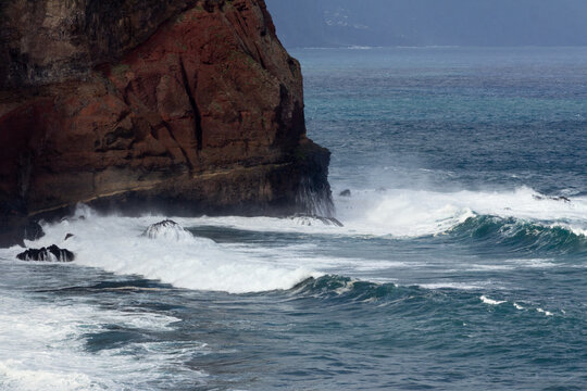 Red rock formations with blue and white waves crashing forcefully against them. The vivid contrast between the vibrant red rock and the dynamic ocean creates a dramatic iimage