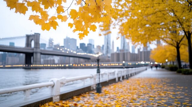 Golden autumn leaves frame a city view. The Brooklyn Bridge is visible in the background, beyond a waterfront promenade strewn with fallen leaves. The image has a shallow depth of field, with soft l