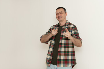 A cheerful man stands confidently in a plaid shirt, smiling while pointing playfully at the camera....