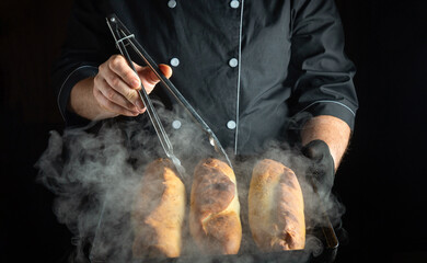 A chef wearing a black coat holds tongs, lifting freshly baked bread from a tray. Steam rises...