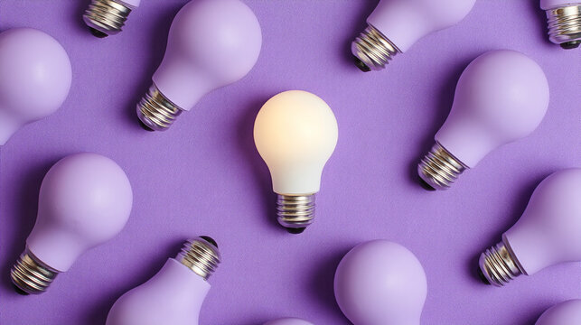 One lit lightbulb among many unlit purple lightbulbs on a textured purple background overhead shot