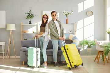 Happy young couple standing in living room with packed suitcases, ready for vacation. Smiling...