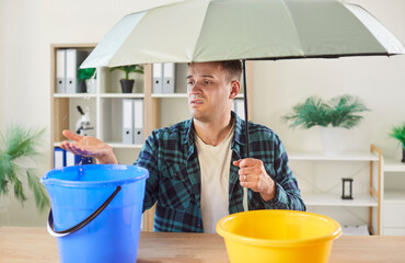 Upset unhappy young man in casual sitting at table in living room at home under umbrella with...