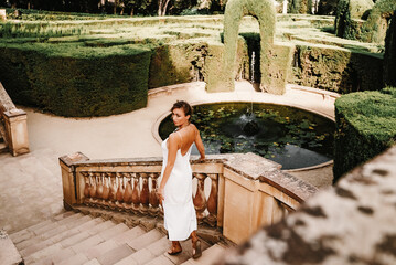 Confident young woman in a white sleeveless dress and gladiator sandals poses beside a pond,...