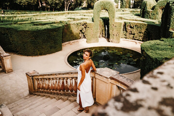 Confident young woman in a white sleeveless dress and gladiator sandals poses beside a pond,...
