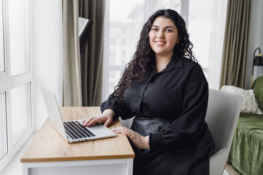 Young pretty caucasian female freelance worker sitting at table desk in her cozy living-room working at laptop online in comfortable conditions, smiling at camera. Advantages of remote job