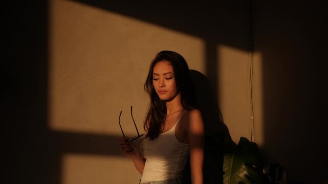 Elegant Asian model holds her glasses and gazes into the warm sunlight. High quality lifestyle and beauty footage perfect for branding, elegance, introspective mood, and fashion promotions.
