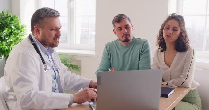 Smiling doctor sitting at desk, discussing medical results with young couple planning pregnancy. Physician using laptop for consultation while patients listening and asking questions in clinic.