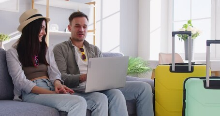 Happy young couple sitting on sofa at home, using laptop and planning trip together. Smiling man and woman surrounded by suitcases selecting tour, booking tickets or arranging vacation details online.