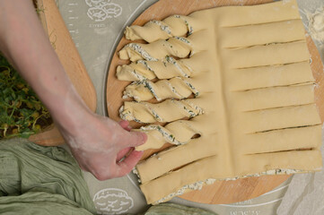 Top view woman's hand twists dough strips on wooden board. Green towel, microgreens sprouts on kitchen table. Home cooking baking puffs