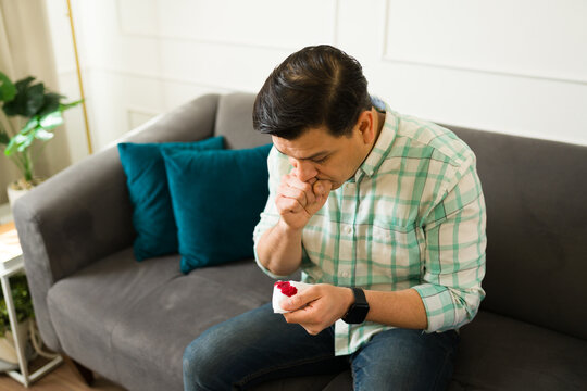 Feverish latino male coughing, wiping bloody sputum while seated on living room sofa during viral illness infection