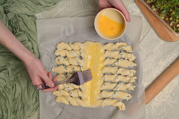 Top view woman's hands smears yolk on the twisted dough strips with a brush. Red small bowl with egg. Rolling pin, green towel, microgreens sprouts on kitchen table. Home cooking baking puffs