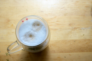 Foamy Cappuccino in Clear Glass Cup on Table
Top view of a cappuccino with thick foam served in a transparent glass mug, resting on a wooden table in natural light.
