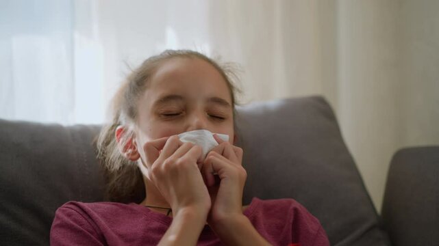 Close-up of little child with red bangle and tied hair sneezing into tissue due to flu, the child appears unwell, lying on the couch with natural light coming through the window