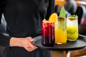 A server presents a dark tray holding three colorful cocktails: a purple drink with citrus, a yellow tropical beverage, and a green kiwi refreshment, perfect for a cheerful gathering.