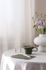 A beautifully serene table display that features delicate flowers in ceramic vase, an elegant green mug, and a charming bag, copybook, candles.