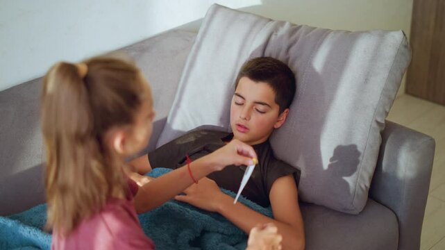 Sister with ponytail and maroon shirt gently removes thermometer from mouth of sick brother lying on couch under blue blanket showing care concern and responsibility