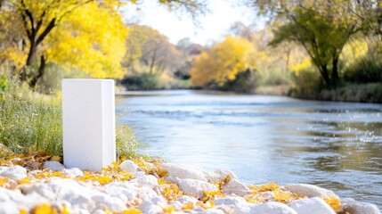 A white rectangular block rests on rocks by a river. Autumn leaves surround it. The background shows a calm river flowing through a vibrant fall landscape. The image is high-quality, brightly lit, a