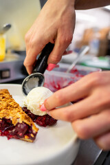 A hand elegantly scoops a generous portion of vanilla ice cream onto a slice of pie adorned with berry compote. The focused preparation highlights the attention to detail in dessert serving.
