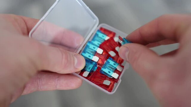 Close-up of hands holding a small plastic box with assorted automotive blade fuses. One blue fuse is being picked up, showing detail and color. Car electronics maintenance concept.
