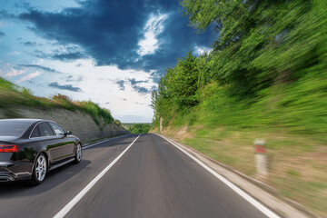 A sleek black car speeds down a scenic countryside road surrounded by lush greenery and dramatic skies, capturing the thrill and freedom of a road trip in motion.