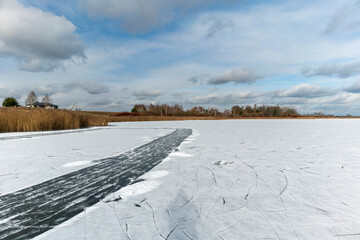 Frozen Lake with Cracked Ice and Winter Landscape. Scenic winter landscape featuring a frozen lake with a prominent crack or ice path cutting through the snow-covered surface. In the distance, dry ree