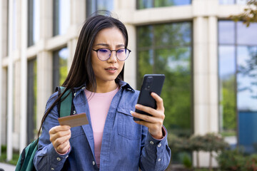 An asian woman is looking at her phone with concern, while holding a credit card in hand, outdoors.