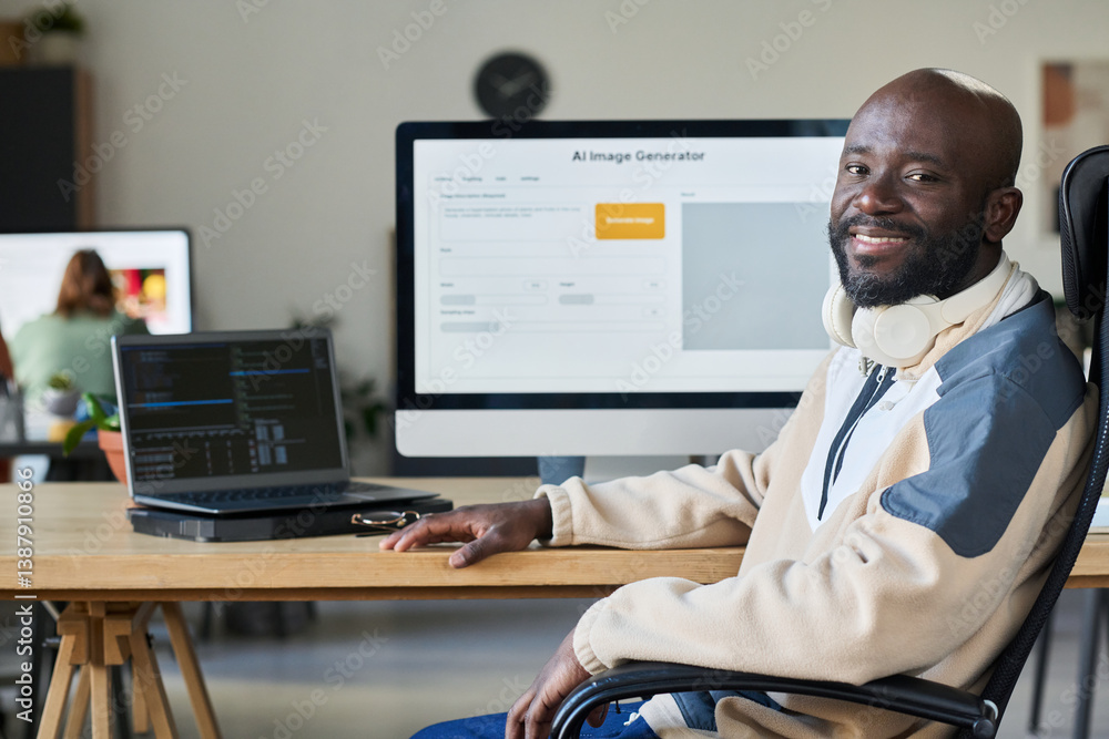 Poster Young successful African American male programmer looking at camera while sitting by workplace with two computers