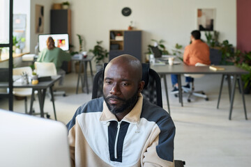 Fototapeta premium Young serious African American programmer sitting by workplace in front of desktop computer and looking at screen