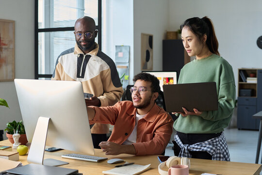 Young woman with laptop and guy with tablet surrounding their colleague making presentation of new software product at meeting - Powered by Adobe