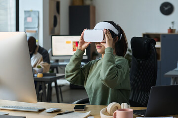 Young female designer in VR helmet sitting by workplace in office and looking through virtual slides during work over project