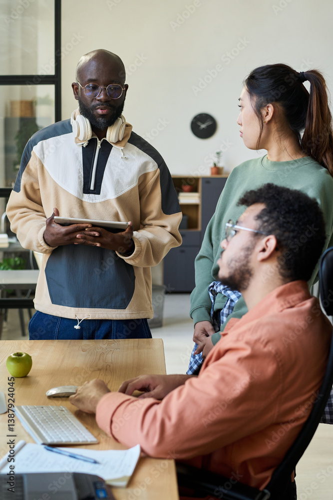 Poster young african american programmer with tablet in hands looking at his colleagues during discussion o