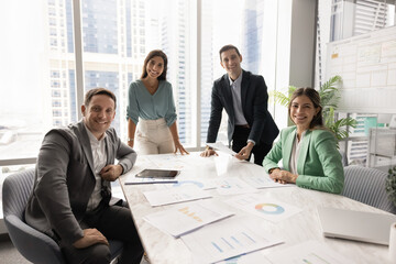 Positive successful business team sitting and standing at large table, looking at camera for group...