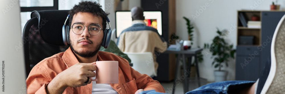 Fototapete header of young restful male office worker in headphones having coffee at break and looking at compu