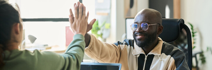 Header of young smiling male programmer giving high five to female colleague sitting in front of him during work over new software