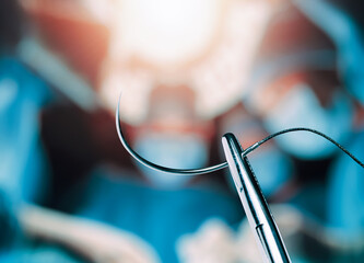 Hemostatic forceps holding half circle cutting needle and suture in front of of blurry operators under the lights in operating room. The surgical team working in an operating room.