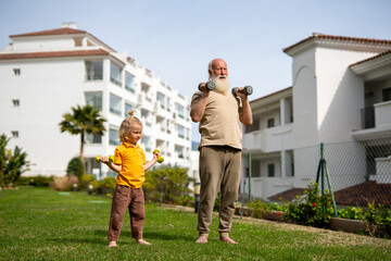 Obraz premium Grandfather and grandson performing shoulder press exercise with dumbbells outdoors