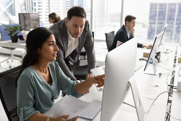Boss and employee discussing work task at computer monitor in open office space. Female Latin...