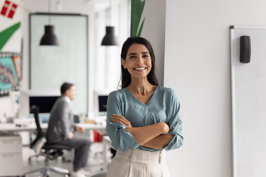 Positive confident beautiful Latin businesswoman posing in office workspace with colleagues working in background, standing with arms folded, looking at camera, smiling