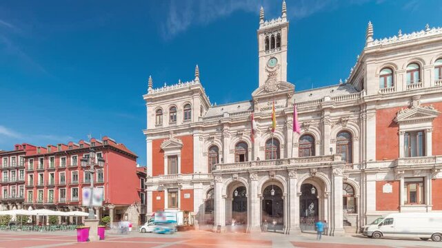 Panorama showing the town hall facade with flags in Plaza Mayor timelapse, Valladolid. Blue sky and historic architecture in the heart of the city. Pedestrians pass by in this central square.