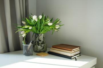 White working desk with notepads, pens and tools and fresh white tulips bouquet on a sunny day