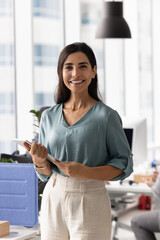 Positive confident Latin businesswoman posing in workspace, holding tablet computer, looking at camera, smiling, standing in office with workplace behind, smiling. Female business leader portrait