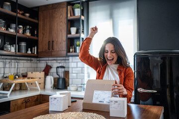 Excited woman unboxing packages in kitchen celebrating online shopping success