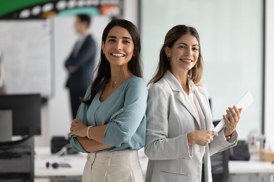 Two positive confident Latin businesswomen posing in office for teamwork portrait. Happy female business colleagues, project partners standing back to back, looking at camera, smiling - Powered by Adobe