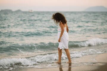 Cute little girl standing on the seashore at the beach near by sea or ocean in summer. Playful...