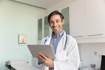 Happy man therapist dressed in white uniform using modern digital tablet device working online, consulting clinic patients remotely, standing alone in contemporary medical office. Technology, workflow