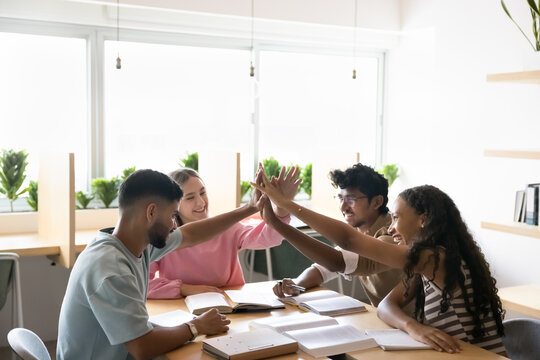 Academic success and collaborative spirit. Happy motivated teen multicultural students peers unite palms in high five above classroom desk celebrate successful teamwork winning grant for group project